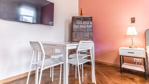 Une pièce avec une table et des chaises blanches, un téléviseur fixé au mur, un grand objet décoratif, une lampe et une table de chevet.