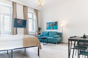 A hotel room with a bed, a sofa, a TV, a dining table, and a painting on the wall.