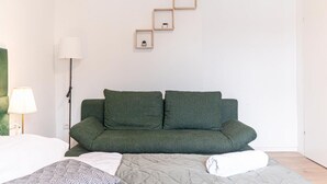 Une chambre moderne avec un canapé vert, un lit recouvert d’une literie à motifs et un luminaire fixé au mur.