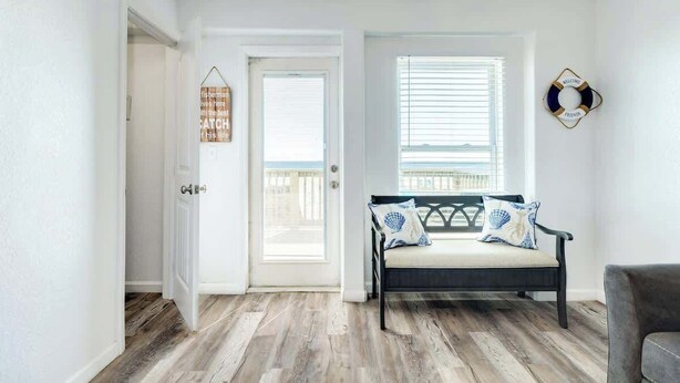 Um quarto com piso de madeira, um banco com almofadas, uma porta e uma janela com persianas.