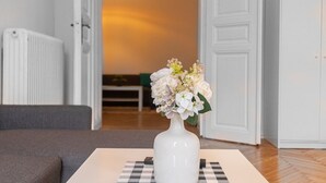 A white vase with flowers on a table in a room with a checkered table runner, a grey sofa, and a white door.