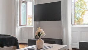 A room with a white table, a vase with flowers, a flat-screen TV, and a window with curtains.