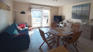 A living room with a sofa, a TV, a dining table with chairs, and a balcony with a view. - Bel Appartement Centre Biscarrosse Plage (BISCARROSSE)
