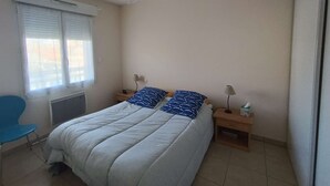 A bedroom with a bed, a ceiling fan, a window with blinds, a chair, and a bedside table with a lamp. - Bel Appartement Centre Biscarrosse Plage (BISCARROSSE)