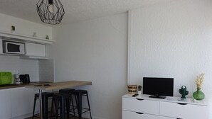 Moderni keittiö, jossa on mikroaaltouuni, kahvinkeitin ja baarituolit. Valkoisen lipaston päällä on televisio ja lähellä pieni pöytä, jossa on kasvi.