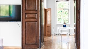 A room with a wooden door, a television, a window with a view of greenery, and a table with chairs.