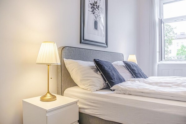 A neatly made bed with white and dark pillows, a bedside lamp, and a framed artwork on the wall.