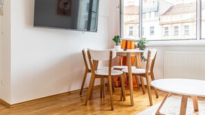 Ett modernt rum med ett matbord, stolar, en platt-TV och utsikt över byggnader genom ett stort fönster.