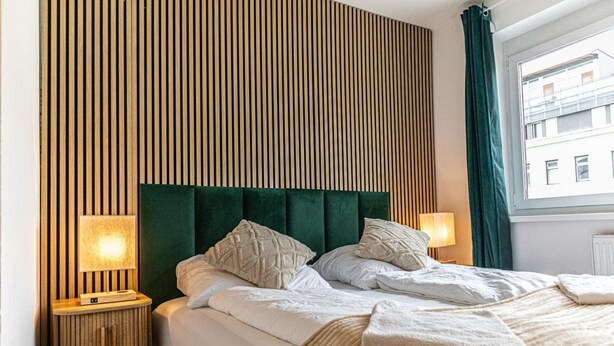 Ett hotellrum med en säng, tv å sänglampor, en vägg med träribbor och ett fönster med gardiner.
