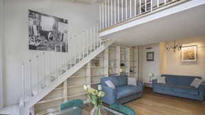 A modern living room with a staircase, blue sofas, a glass coffee table, and a bookshelf. - Florentine Gem Apartment (Firenze)