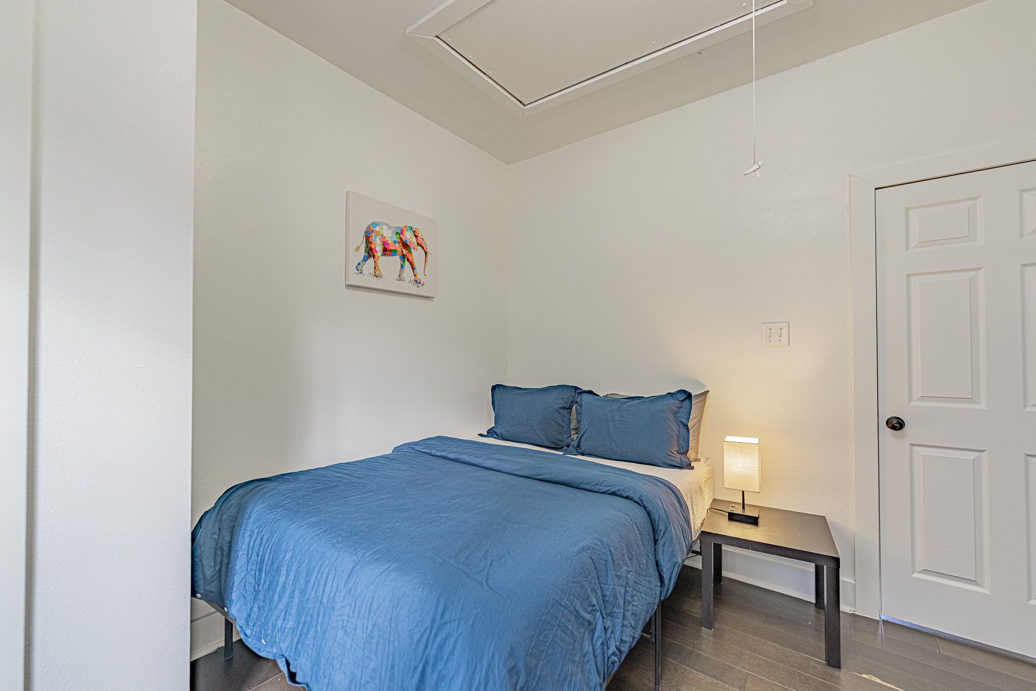 Ein Schlafzimmer mit einem Bett, einem Nachttisch mit Lampe, einer Tür und einem Wandbild.