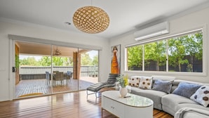 A spacious living room with a wooden floor, a white sofa, a glass coffee table, and a large window with a view of the outdoors.