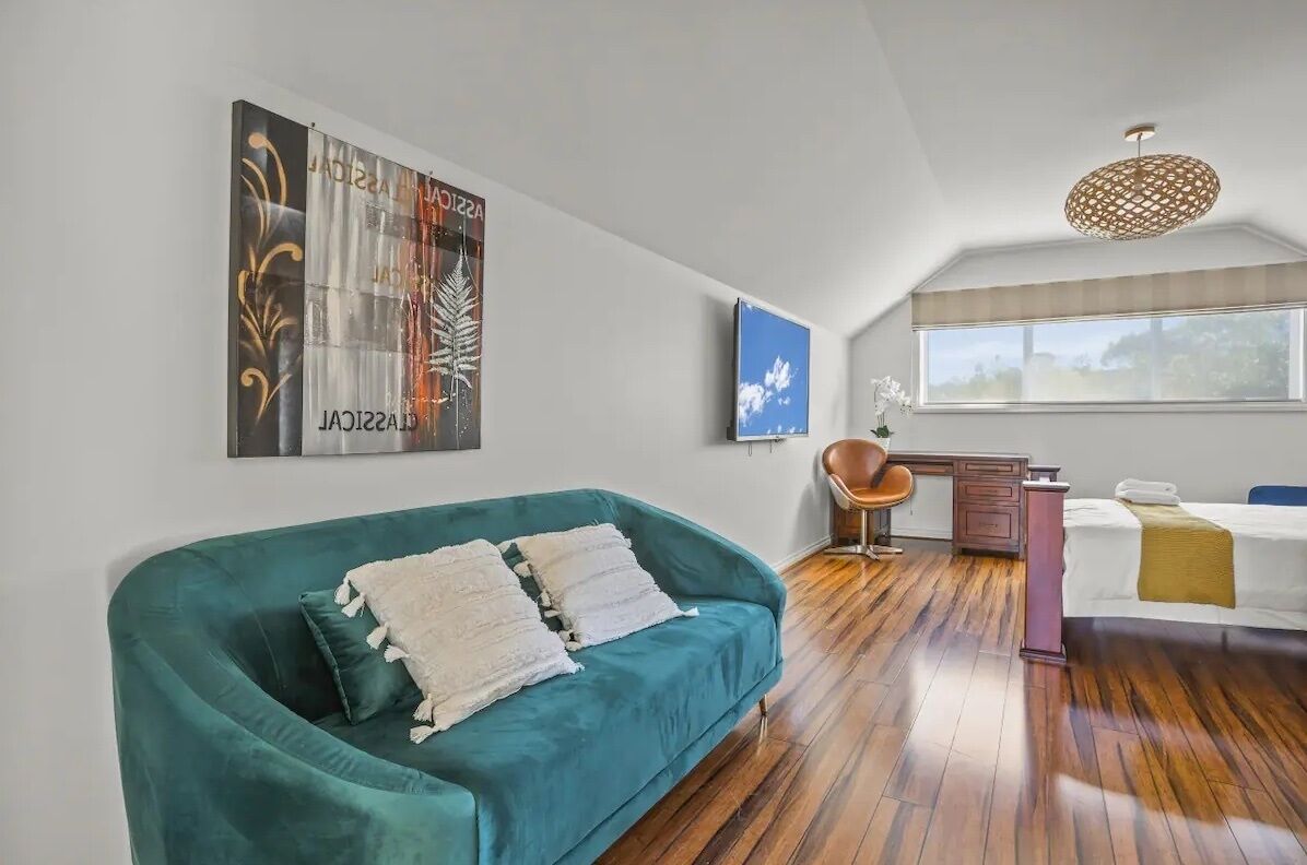 A modern living room with a teal sofa, a wooden floor, a desk, and a bed.