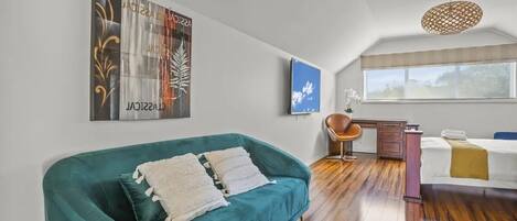 A modern living room with a teal sofa, a wooden floor, a desk, and a bed.