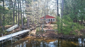 A cabin with a red roof surrounded by a forest, a dock extending into a pond, and a small bridge connecting the dock to the cabin.