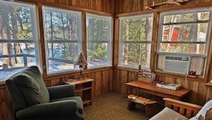 A cozy room with a green armchair, a beige sofa, a wooden table with books, and a lamp. Large windows with blinds offer a view of trees and a red building outside.