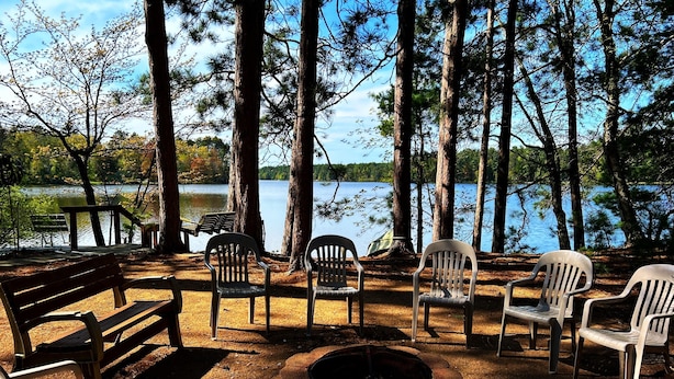 A campsite with a fire pit, wooden benches, and plastic chairs around a lake.