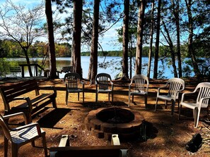 Un camping con un hoyo para fuego, bancos de madera y sillas de plástico alrededor de un lago.