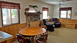 A cozy living room with a wooden dining table, leather chairs, a stone fireplace, and a ceiling fan.