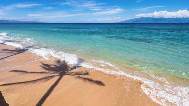 Une plage à l’eau turquoise et limpide, un rivage sablonneux et une montagne qui se profile à l’horizon.