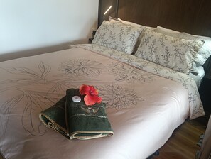 Cama bem arrumada com roupa de cama floral, toalha verde com flor vermelha e um pequeno objeto decorativo no travesseiro.