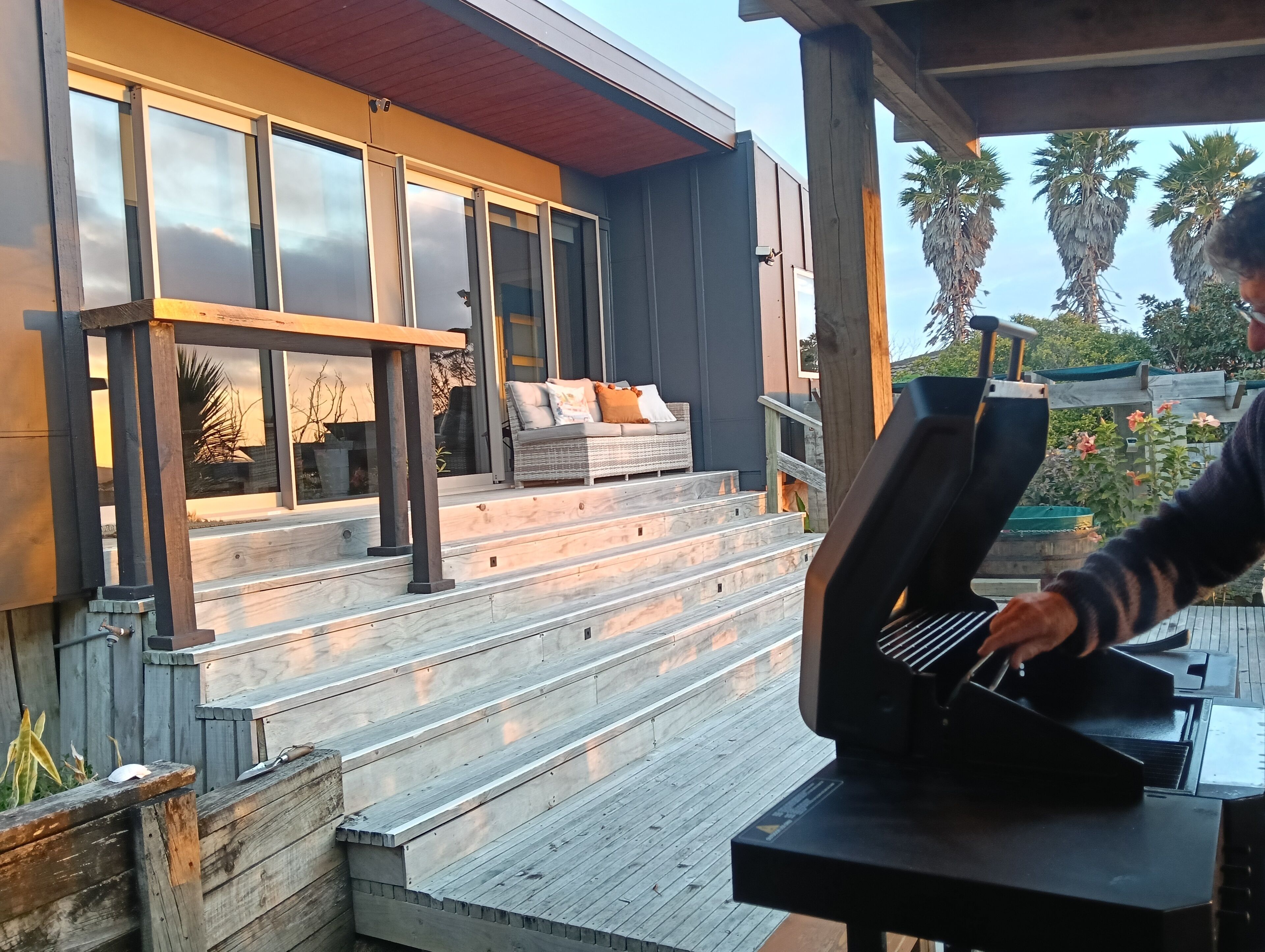 A modern outdoor kitchen with a built-in grill, wooden deck, and a seating area with a sofa and cushions.