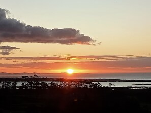Auringonlasku rannikkomaisemassa, jossa puiden ja kukkuloiden siluetit näkyvät etualalla.