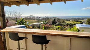 Terraza de madera con zona de bar que incluye dos taburetes negros y vistas a un paisaje con colinas y una masa de agua.