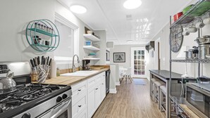 Une cuisine moderne avec des armoires blanches, un plan de travail en bois et des appareils électroménagers en acier inoxydable. On y trouve des étagères remplies d’ustensiles de cuisine et une horloge au mur.