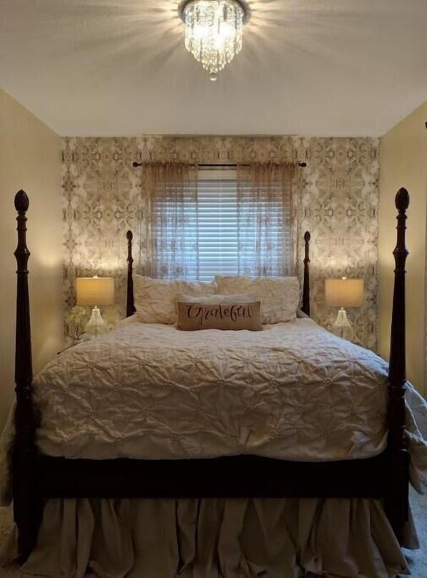 Een slaapkamer met een hemelbed, een kroonluchter, behang met een patroon en een raam met luxaflex.