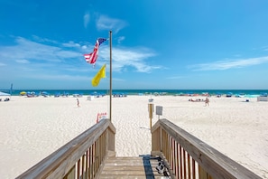 Une promenade en bois menant à une plage où l’on peut voir des drapeaux, des parasols et des personnes se prélassant au soleil.