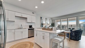 A modern kitchen with white cabinets, a central island, and a dining area with a blue chair and a white stool. - DIRECT BEACH ACCESS, GULF VIEWS, ZERO-ENTRY POOL! (Gulf Shores)