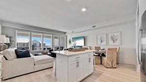 A modern living room with a sofa, a dining area with chairs, a kitchen island, and a large window with blinds. - DIRECT BEACH ACCESS, GULF VIEWS, ZERO-ENTRY POOL! (Gulf Shores)