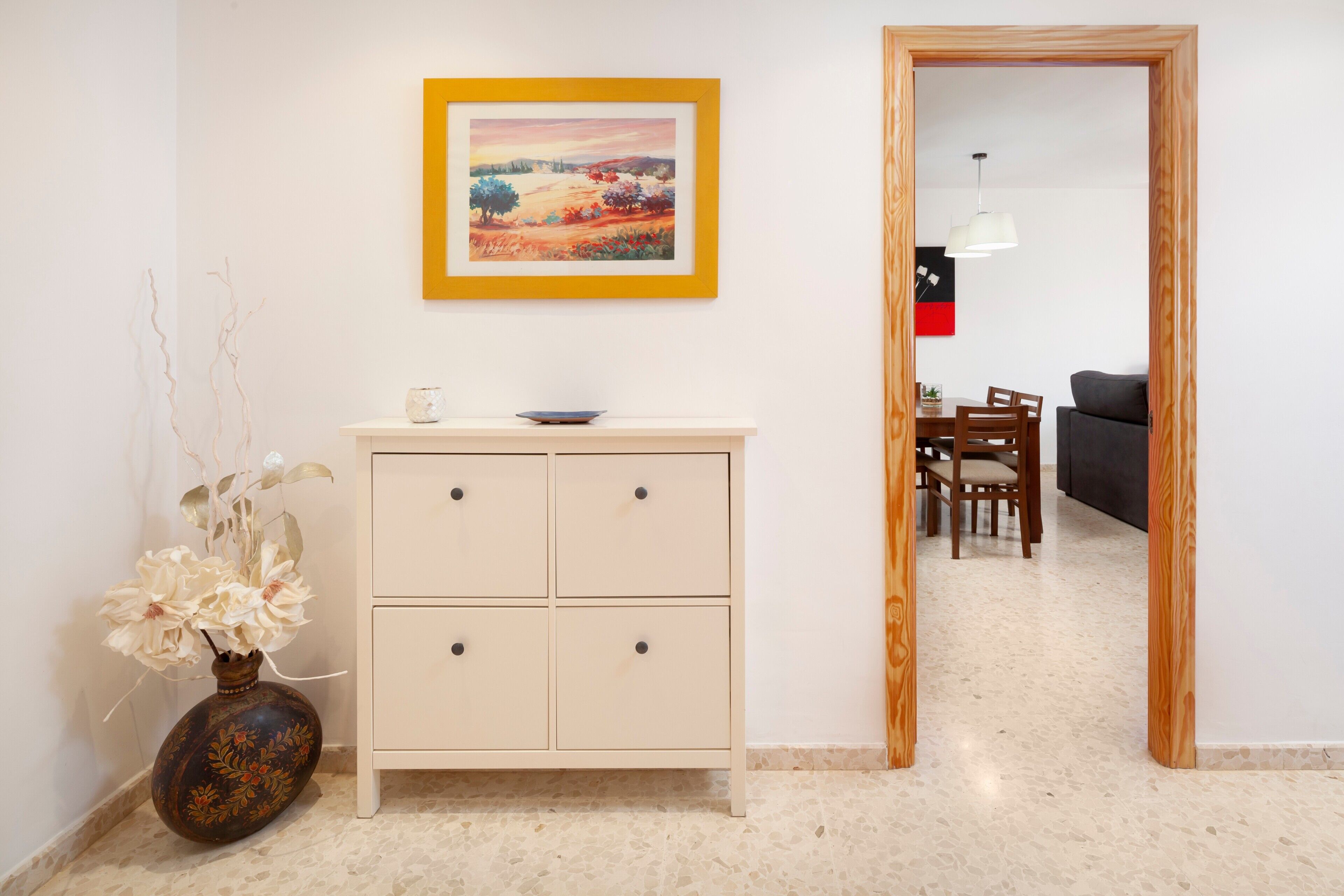 Een kamer met een witte kast, een vaas met bloemen, een ingelijst schilderij en een eethoek met stoelen en een tafel.