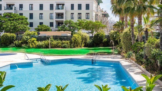 A swimming pool with steps leading into it, surrounded by greenery and a building with balconies.