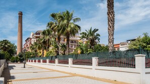 A paved walkway, a tall chimney, palm trees, and a building with balconies.