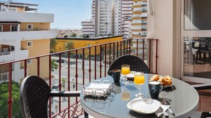 A balcony with a table set for breakfast, including croissants and orange juice, overlooking a cityscape with buildings.