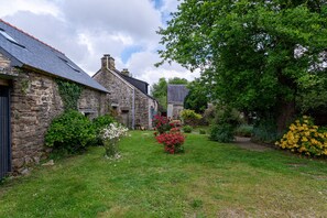 Un bâtiment en pierre doté d’un toit en pente, d’une cour verdoyante et de diverses plantes à fleurs.
