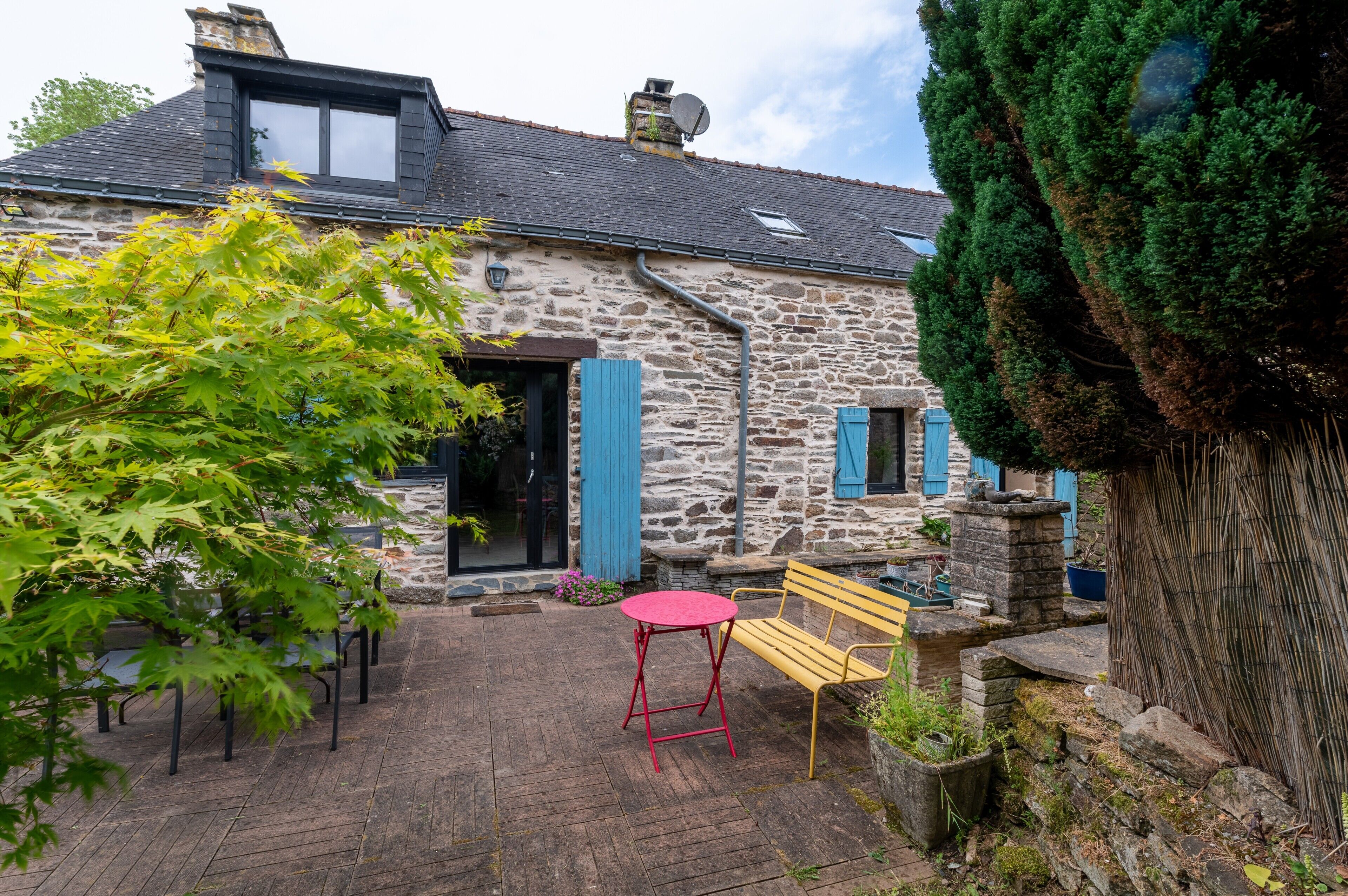 A stone building with a sloped roof, a red table, and a wooden bench in the courtyard.