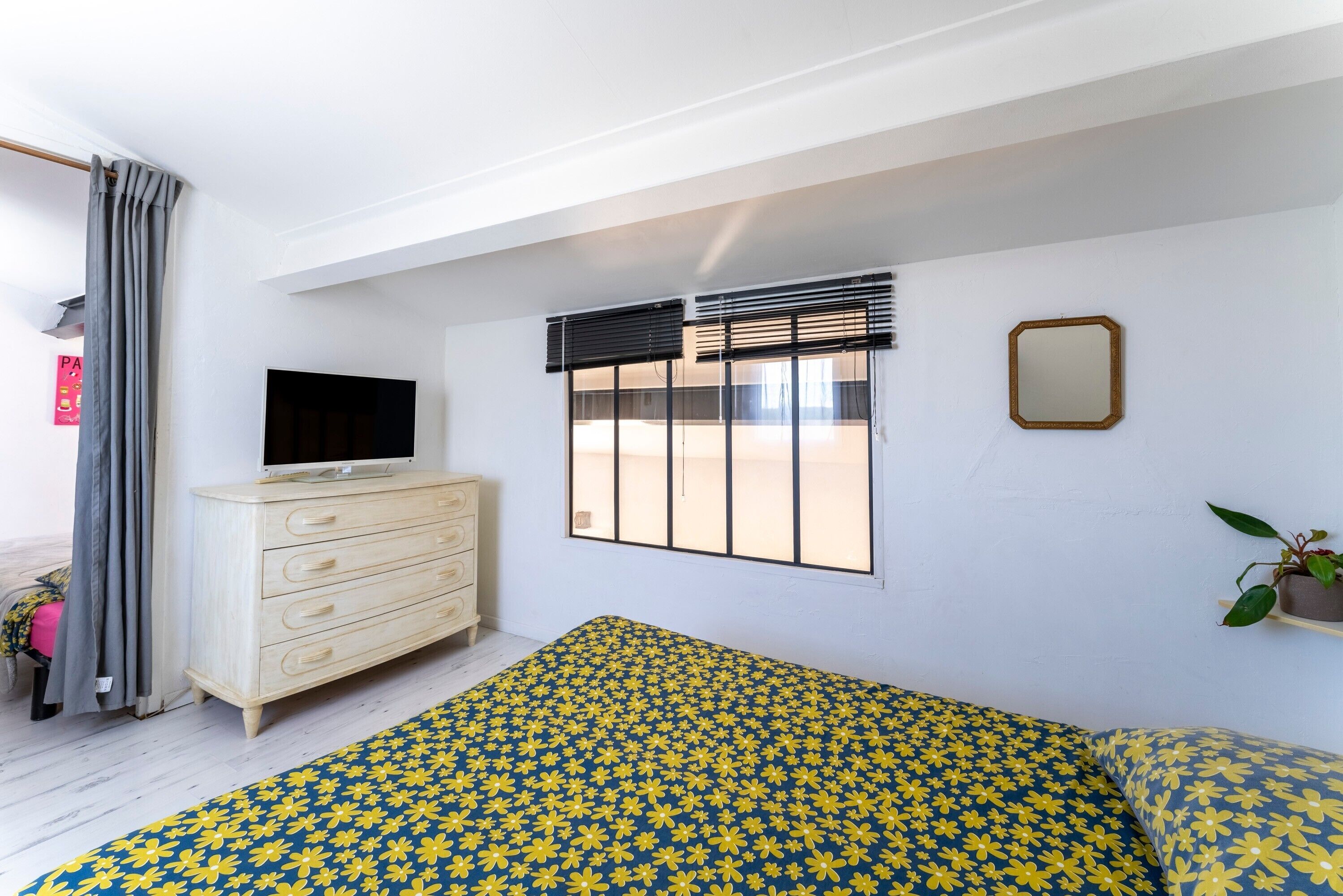 Une chambre à coucher avec un lit, une commode, un téléviseur, une fenêtre avec des stores et une plante en pot.