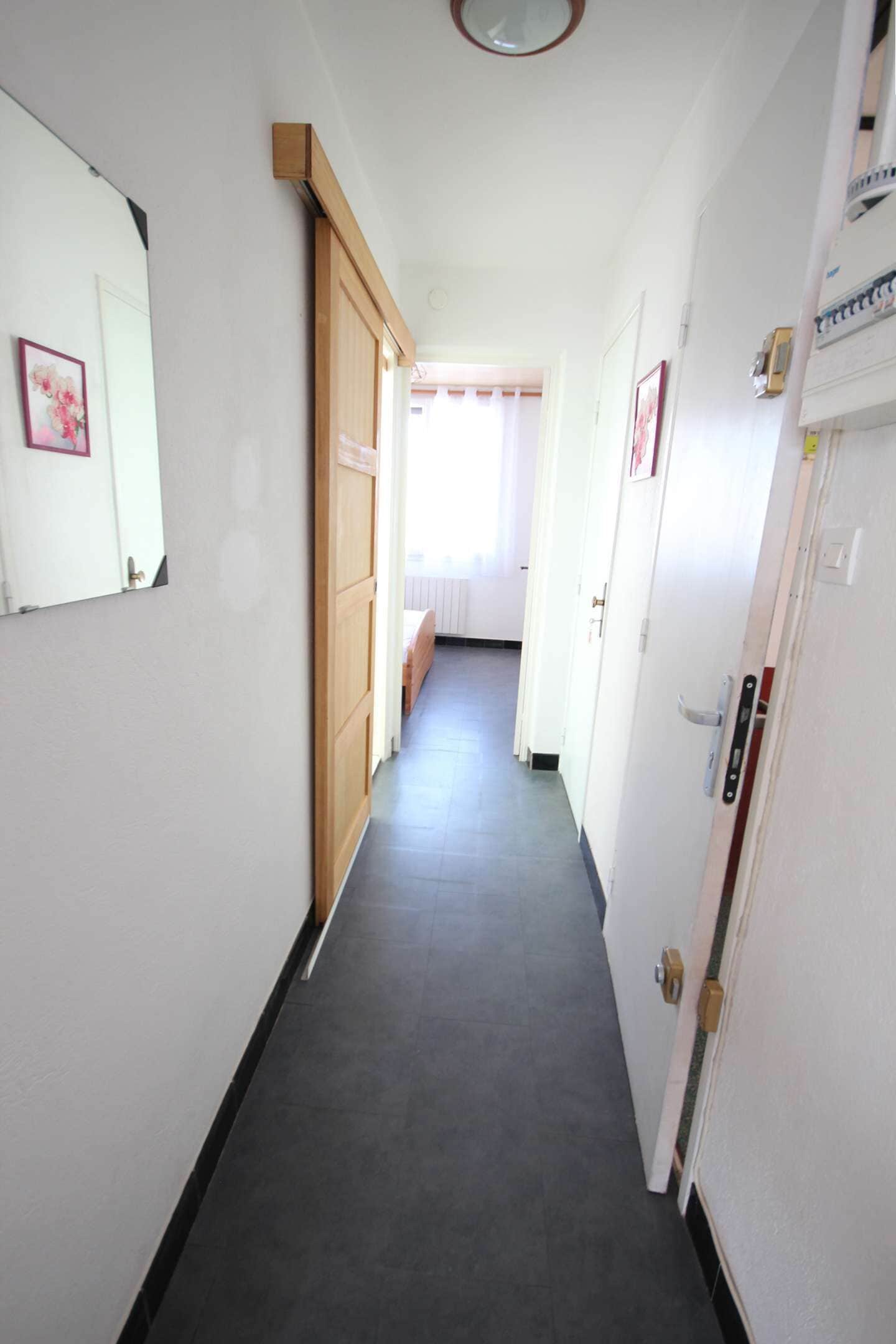 A narrow hallway with a wooden door on the left, a white door on the right, and a bench in the distance.