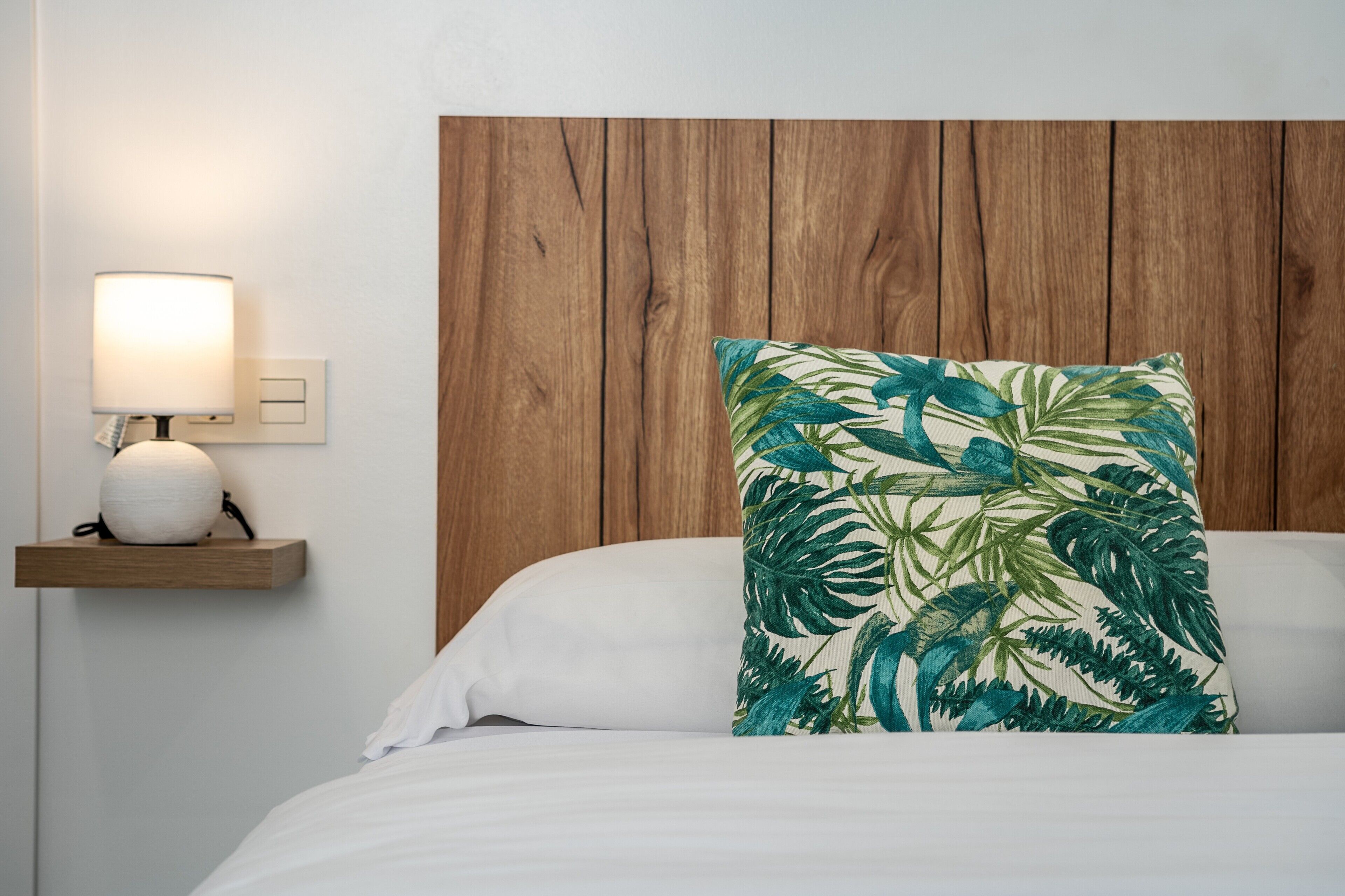 A bed with a white pillow and a tropical leaf-patterned pillow, a bedside table with a lamp, and a wooden headboard.