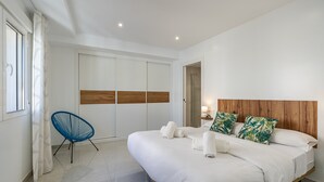A modern bedroom with a large bed, a blue chair, a wardrobe, and a shower area.