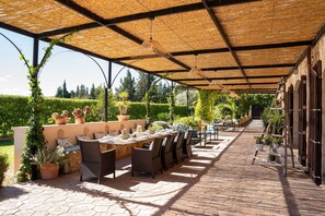 Un patio al aire libre con zona de estar cubierta, mesas y sillas, rodeado de plantas en macetas y un seto.