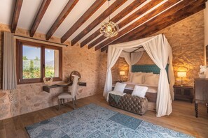 Un dormitorio con pared de piedra, vigas de madera, cama con dosel, un escritorio y vistas por la ventana.