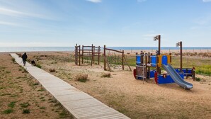 A beach with a wooden walkway, a playground with a slide, and a person walking a dog.