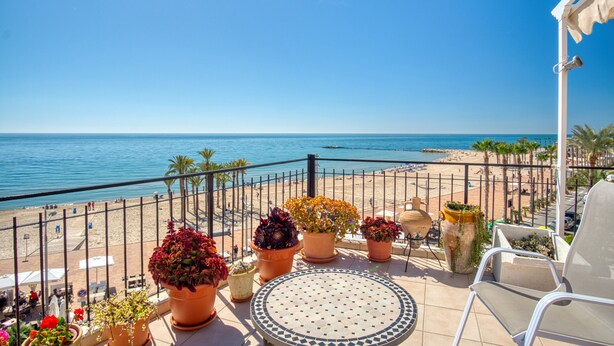 Ein Balkon mit Topfpflanzen, einem runden Tisch und Blick auf Strand und Meer.