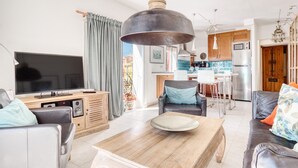 Ein modernes Wohnzimmer mit Fernseher, Sofa, Couchtisch und Essbereich, im Hintergrund ist eine Küche zu sehen.