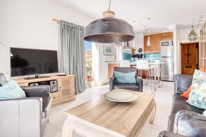 Un salon moderne avec un téléviseur, un canapé, une table basse et un coin repas, avec une cuisine en arrière-plan.