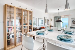 Un coin repas avec une table blanche dressée pour quatre personnes, entourée de chaises. On y trouve également un meuble en bois avec des portes en verre, contenant des livres et des objets décoratifs.